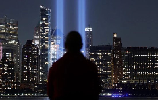 From the back, a silhoetted man stares at a nighttime city skyscape as two large blue beams of light reach to the sky