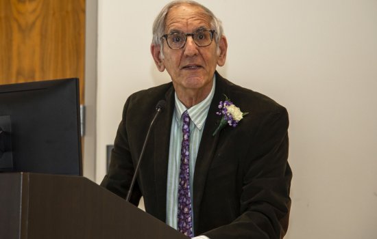 Professor Berman at a podium, wearing glasses, a brown jacket and purple tie, and a flower in his lapel.