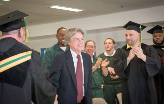 Randal Craig, smiling in a blue suit and red tie, is surrounded by clapping men in graduation garb or prison garb