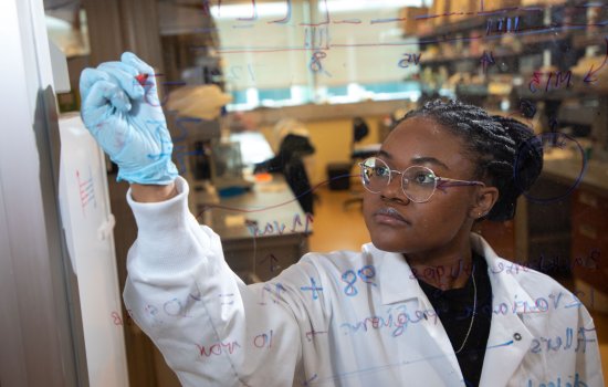 A woman in a lab coat and blue goves is seen behind a pane of glass she is writing formulas on.