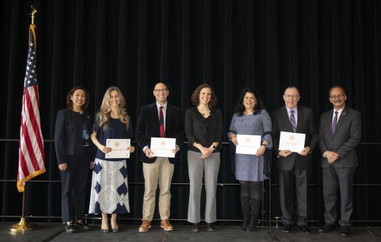 Seven smiling people, some holding awards certificate, stand in a row on a stage next to an American flag