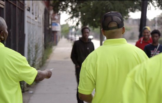 Three men in yellow shirts, their backs to the camera, approach three young men on the street.