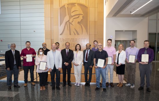 A group of faculty stand in front of the UAlbany Minerva logo in the ETEC building with recognition awards in hand.