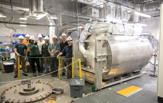 A group of scientists and movers stand near a large cylinder dark matter tank inside of a lab room.