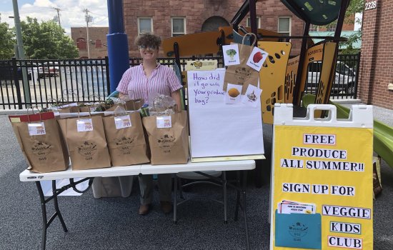 Melissa Toback stands outside behind a table that holds several large brown bags filled with produce.