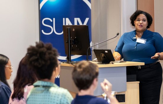 A woman speaks at a podium indoors in front of a seated crowd with a SUNY logo behind her.