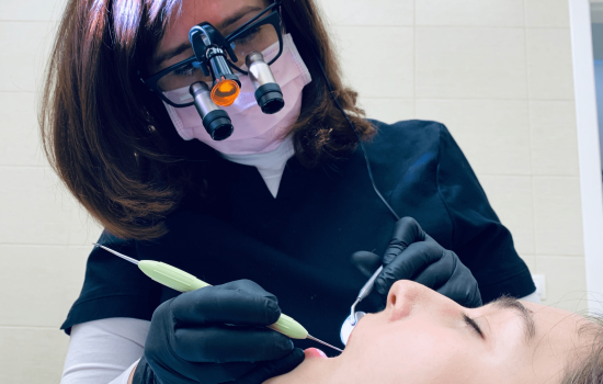 Image of a dental cleaning: A woman with dark brown hair wearing dark-rimmed glasses with magnifying attachments, wearing a paper face mask and latex gloves, cleans the teeth of a reclining person in the foreground using metal dental tools.