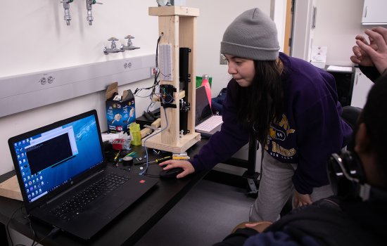 A young woman works on laptop while standing indoors with an engineering project in the background.