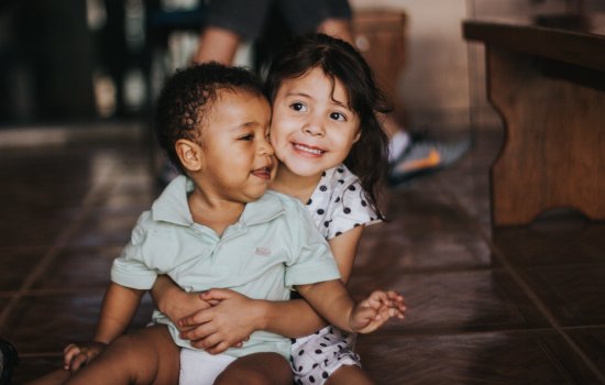 A small child is sitting on the floor, hugging a toddler who sits on her lap.