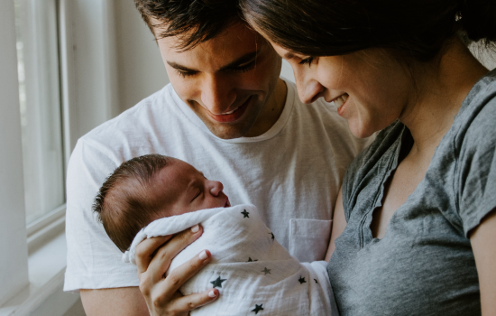 A man and a woman are holding a baby swaddled in a blanket and are smiling at the baby.