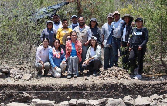 Verónica Pérez Rodríguez poses with members of the Cerro Jazmin Archaeological Project crew in Oaxaca, Mexico, in front of an archaeological excavation site populated with trees, dirt and rocks.
