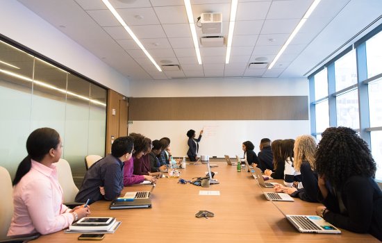 A group of professionals sit at a boardroom style table watching an African American woman at the white board giving a presentation.
