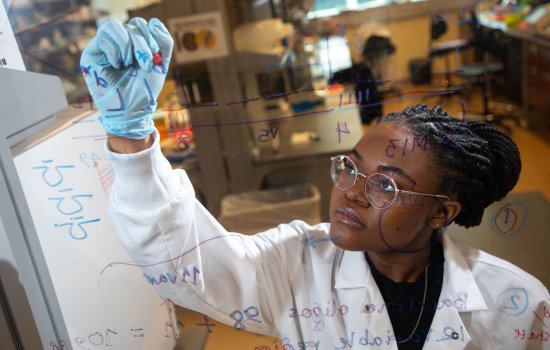 A student in a white lab coat is photographed through a glass window as she reaches up to write equations on the glass in purple and blue ink.