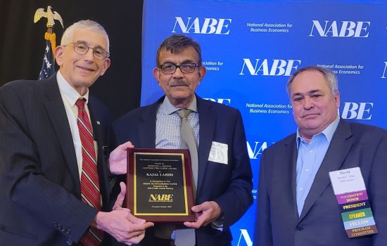 Three men in suits stand in front of a blue screen with the initials NABE printed several times. The man in the center is being handed a plaque on which his name, Kajal Lahiri, can be read.