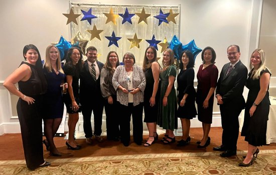 A group of 12 smiling people wearing formal attire stand in a row in front of a gauzy draped backdrop decorated with twinkle lights, gold and blue cutout stars, and gold and blue star shaped balloons.