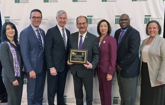 A group of seven people stand in front of a screen at the Association of Public Land-Grand Universities holding a plaque naming UAlbany as an Innovation and Economic Prosperity University.