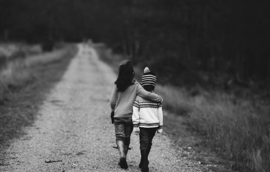 Two young children walk together down a wooded path; the taller child has their arm around the smaller one. 