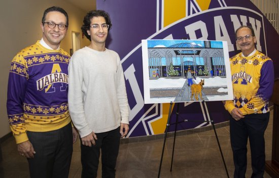A student in a white shirt stands by a large image of his greeting card illustation, flanked by men wearing UAlbany shirts with snowflack designs