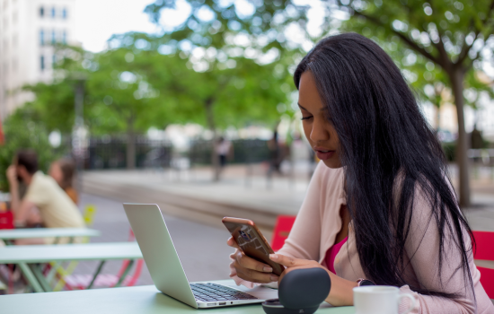 A woman sits at an outdoor table, typing on her phone while on her laptop.
