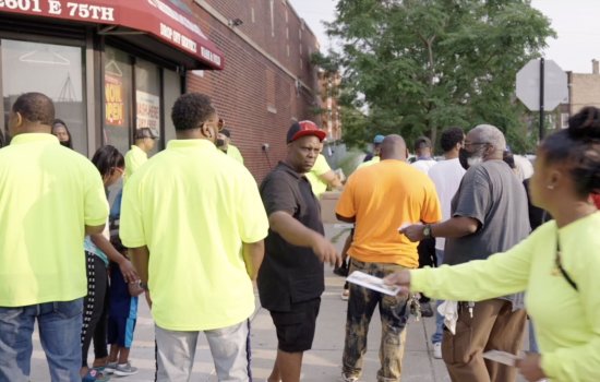 A group of people on a sidewalk. A woman in a yellow shirt hands a man a pamphlet as other men in yellow shirts stand nearby.