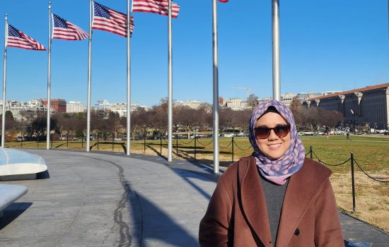 A smiling woman in a headscarf and winter coat stands on a path with flagpoles flying the American flag