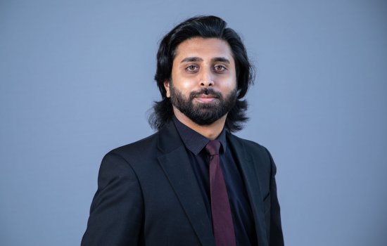 Portrait of a man with shoulder-length black hair and black beard, wearing a black suit jacket and purple tie, against a blue background.