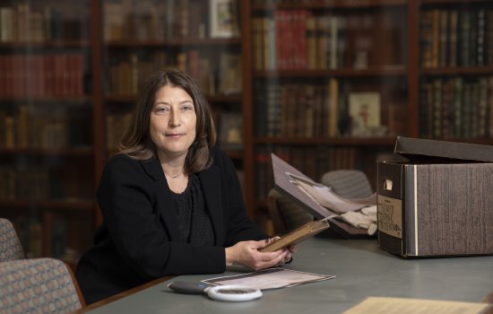 A woman with short brown hair dressed in black sits at a desk holding a gold leaf-trimmed book, a box of papers and a magnifying glass in front of her, and a glass case of books behind her.