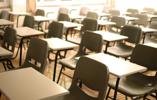 Rows of white plastic desks with gray plastic chairs are arranged in a classroom. No student are present. Sunlight is streaming through the windows. 