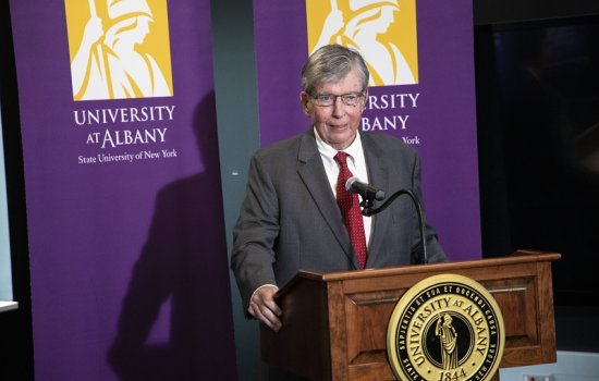 NYS Sen. Neil D. Breslin stands wearing a dark gray suit and red tie speaking at wooden lectern with the UAlbany seal framed on either side behind him by two purple UAlbany banners with the University's gold Minerva logo.