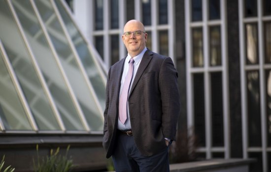 A man in a suit jacket and tie stands near glass windows