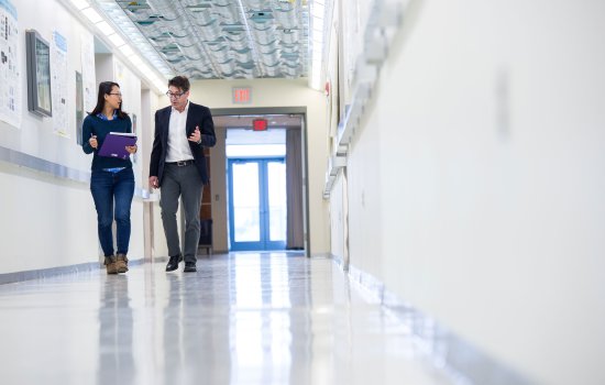 A woman wearing glassesholding a binder and pen walks forward with a man wearing glasses and a jacket in a hallway.