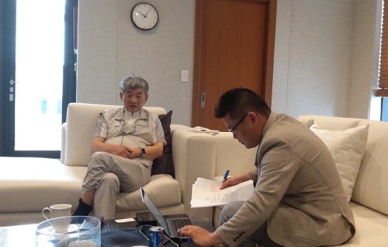 An older man with silver hair is seated in a chair next to a younger man with black hair and glasses, who sits on a couch nearby consulting a handful of papers and looking at a laptop. They are gathered around a table with a pitcher of water and other beverages.