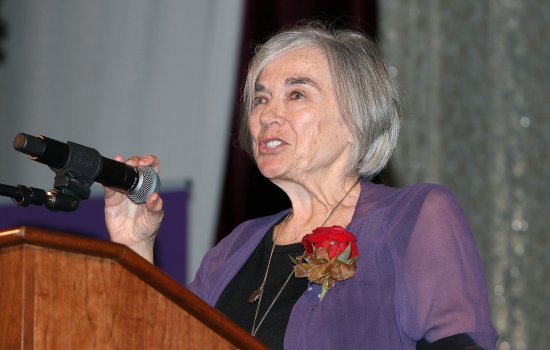 A woman with short gray hair wears a purple blouse and rose lapel as she stands at a podium with a hand grasping a microphone