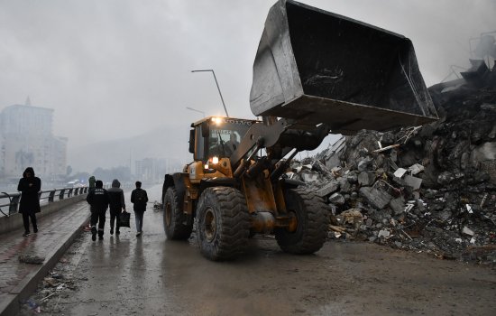 A bulldozer lifts its blade high into the air next to a pile of rubble along a pier as people dressed in black walk by. The sky is gray and hazy and the street is littered with debris.
