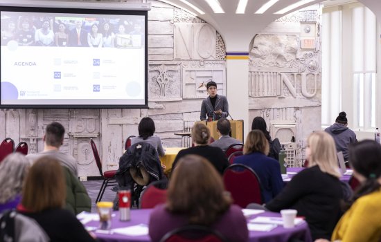 A woman speaks from a podium near a screen with a projected agenda on it. The backs of heads of the audience, seated at round tables, is seen in the foreground.