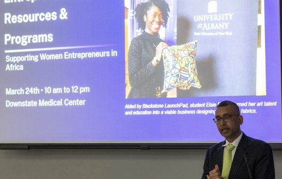 A man in a green tie speaks at a lectern with a presentation screen behind him featuring Blackstone Launchpad UAlbany student Elsie Essien.