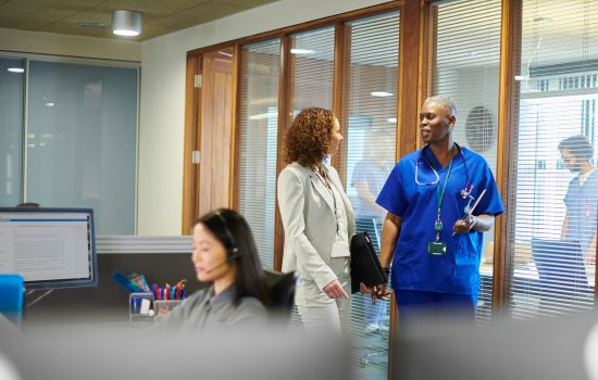 Three people - two standing and speaking to each other, one sitting at a computer wearing a headset - are pictured indoors in a healthcare setting.