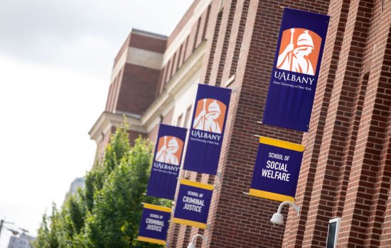 UAlbany's School of Social Welfare building on the Downtown Campus. The building is red brick, with three sets of purple and gold UAlbany flags mounted on the side. Each set of flags has the Minerva logo on top. Then below, one flag says School of Social Welfare; the other two say School of Criminal Justice.