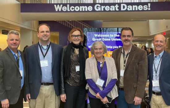 Six people in business attire and lanyards pose in the UAlbany Student Union. Behind them is a digital sign that reads "Welcome to the Great Dane Union", and "Welcome Great Danes" is painted on the wall above.