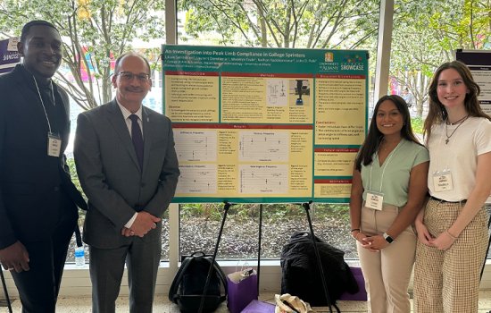 Students, with UAlbany President Havidán Rodríguez stand on either side of a presentation board titled, "An Investigation into Peak Limb Compliance in College Sprinters." President Rodríguez is wearing a gray suit and the students are dressed professionally and wearing lanyards.