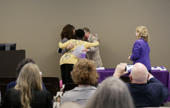 Students huddle around a teacher for a group hug near a purple table as an audience looks on.