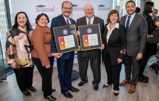Six smiling people face the camera in a line, the two men in the center holding framed award certificates,