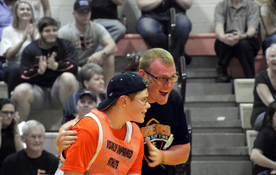 A student athlete celebrates with his visually impaired teammate after he scores in basketball as fans cheer from the stands.