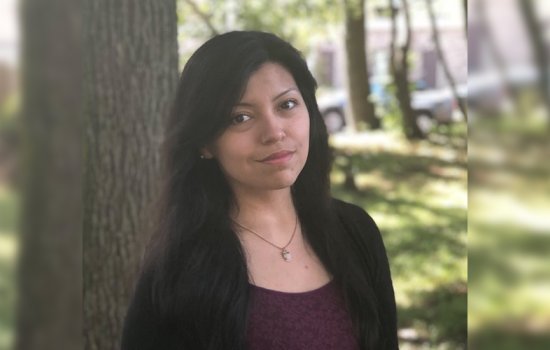 A woman with dark hair, a maroon top and a black cardigan smiles at the camera. She stands in front of a tree. Grass, sunlight and a couple of cars are in the background.