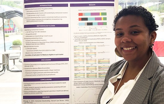 Alyssa Kamara smiles in front of a poster. On the poster, headings of white text in purple boxes organize the information. The headings read: "Introduction," "Methodology & Data," "Discussion," "Conclusions," "Sources," and "Results."