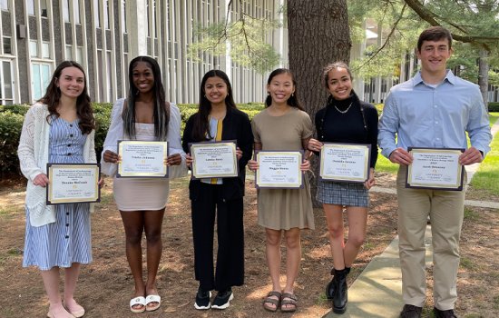 Six students stand outside on campus on a sunny day while holding award certificates.