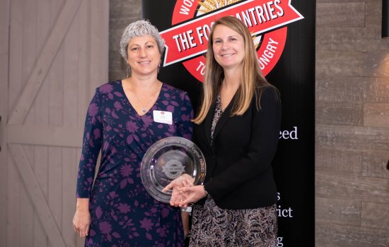 Janine and Natasha hold a circular glass award and stand in front of a sign that says The Food Pantries.