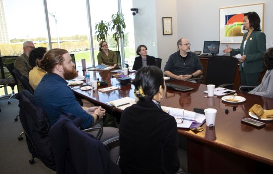 A woman in a green suit speaks to faculty seated around a table in a conference room.