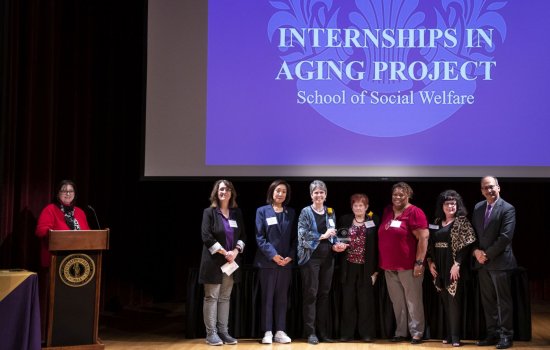A woman stands at a podium near a line of seven smiling people standing under a screen with the words "Internships in Aging Project/ School of Social Welfare"