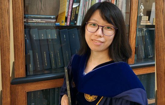 A women wearing glasses and a blue velvet PhD hood stands in front of a bookcase